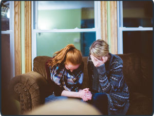 Zwei Frauen beten und weinen gemeinsam auf einem Sofa – Symbol für Seelsorge, Trost und geistliche Begleitung