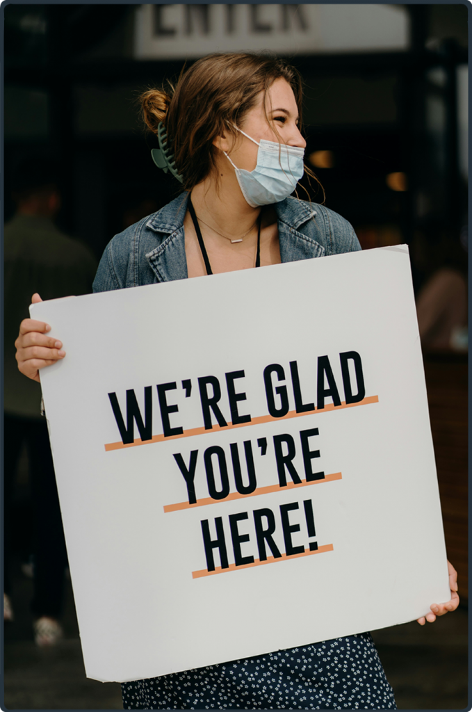 Eine junge Mitarbeiterin einer Gemeinde hält ein Schild mit ‚We’re glad you’re here!‘